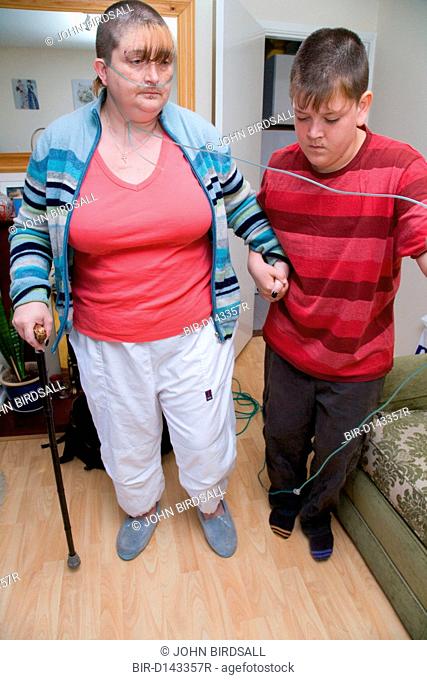 Young carer helping his mother, who is severely asthmatic, to walk across room whilst holding her oxygen tubes