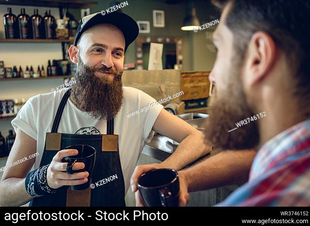 Dedicated hairstylist with hipster beard smiling while drinking coffee with his male customer and friend before haircut in the interior of a trendy hair salon