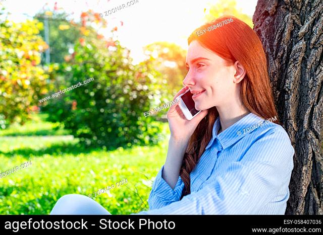 Young redhead woman talking on smartphone under tree in park on sunny day. Beautiful girl relaxing outdoors. Charming girl sitting on green grass leaning...
