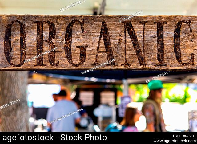A close up view of a rustic wooden sign with the word organic, hanging above blurry people as they shop at a local farmers market