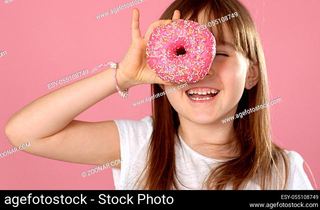 Young beautiful happy and excited blond girl 6 or 7 years old holding donut on her eyes looking through. Playing cheerful with cute smile while posing over ping...
