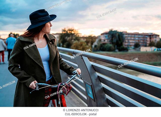 Young woman with bicycle looking out from footbridge at dusk, Florence, Tuscany, Italy
