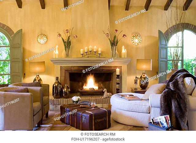 Old luggage trunk & brown armchairs in Spanish country living room with fire lit in fireplace & faux fur throw on sofa