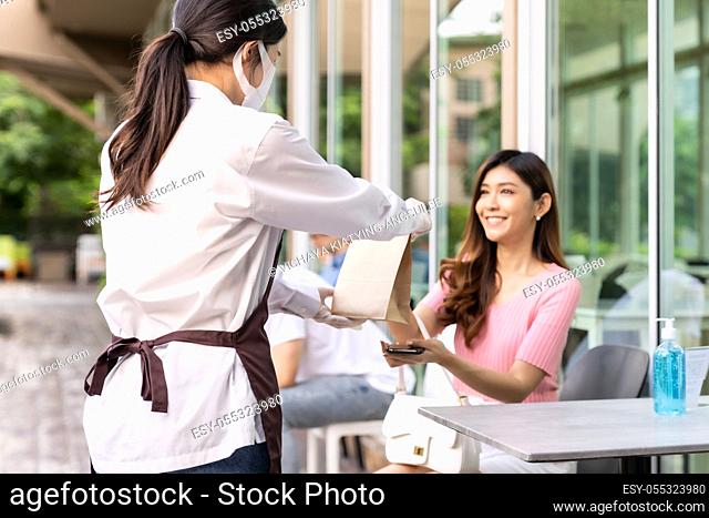 Back view of asian waitress with face mask give order of take out food bag to attractive woman female customer. Take away or take-out food service concept in...