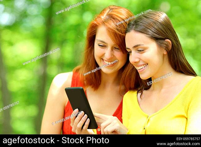 Two happy friends checking smart phone standing in a green park