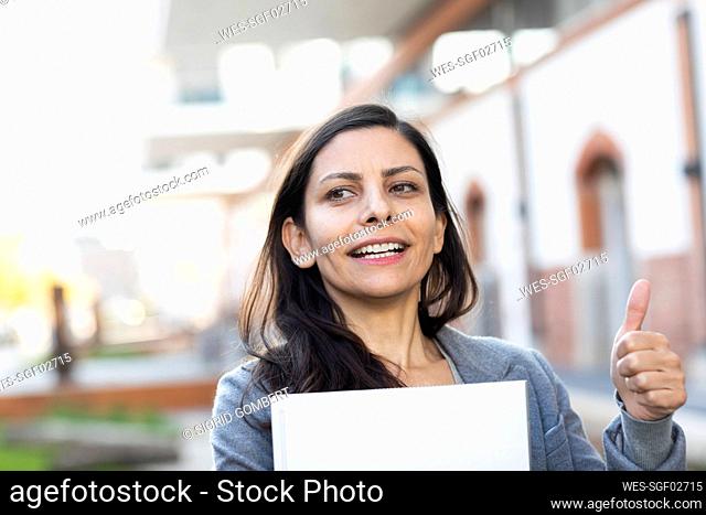 Businesswoman with laptop showing thumps up while standing against building
