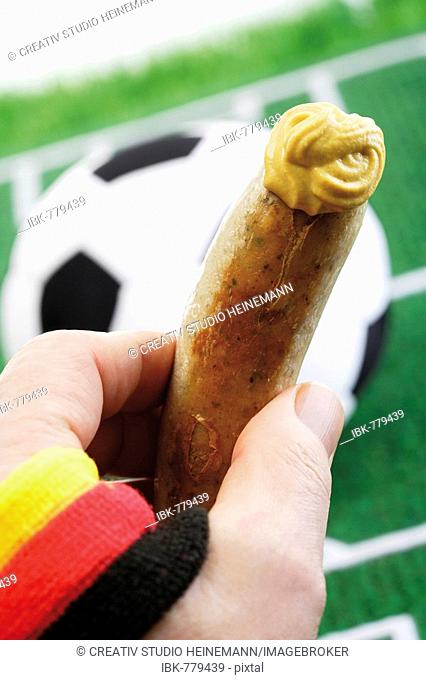 Hand holding bratwurst sausage with mustard in front of a football field with a soccer ball, German national colours
