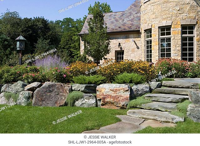 PATIOS: Backyard of French Normandy home, limestone patio and steps, close up, stone steps lead to house on right, large rocks terrace a perennial flower border