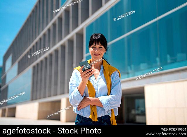 Smiling mature businesswoman with smart phone in front of building