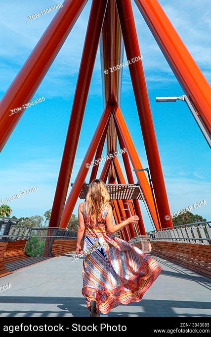 Flamboyant woman in long sundress of vibrant colours and pattern is walking across Yandhai Nepean Crossing, a new bridge with Darug aboriginal name meaning...