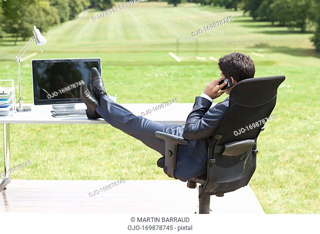 Businessman with feet up sitting at desk in field talking on cell phone