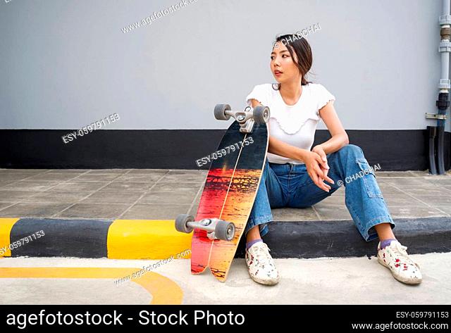 Young asian woman with sunglasses on her head resting after skateboarding. The skateboard is placed on the edge of the pedestrian lane