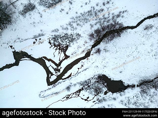 29 November 2023, Hesse, Schmitten: A stream meanders through the snow-covered winter landscape in the Taunus (aerial photo taken with a drone)