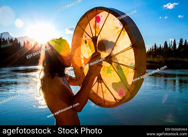 A creative back lit into the sun shot with lens flare, of a spiritual guy playing traditional native music with a handheld sacred drum by tranquil lake