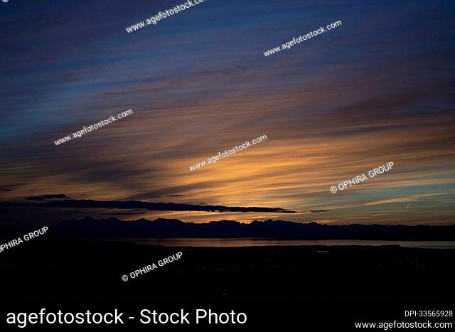 The sun sets above the Tordrillo Mountains across Cook Inlet from Anchorage, Alaska, on Dena’ina land, with the Knik arm in view; Alaska
