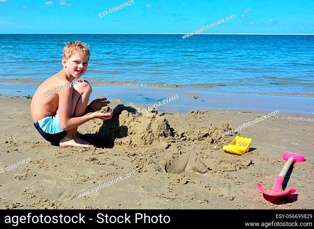 Little blond boy with swimming trunks builds a sand castle in the sand on the beach in front of the sea, with a blue sky