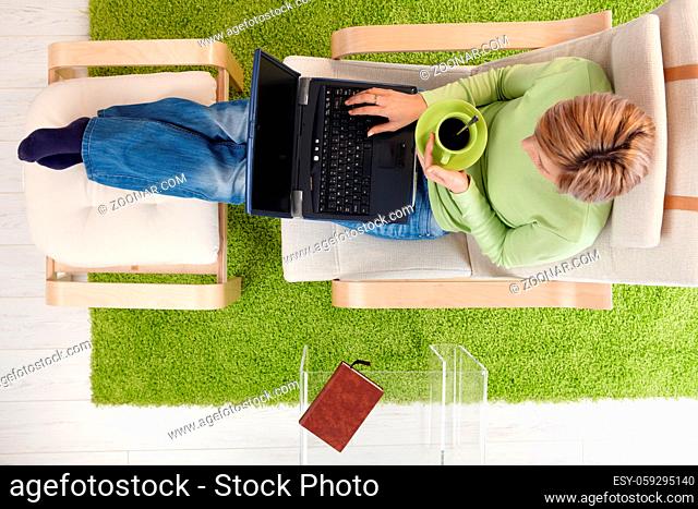 Woman using laptop, drinking coffee, sitting in armchair with legs up on footboard in overhead view