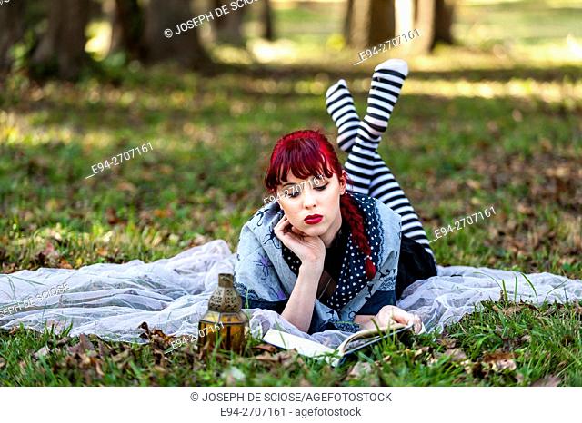 A pretty 23 year old red headed woman outdoors, lying on the grass looking down at a book