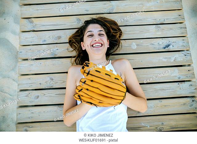 Young woman lying on wooden path laughing with baseball glove