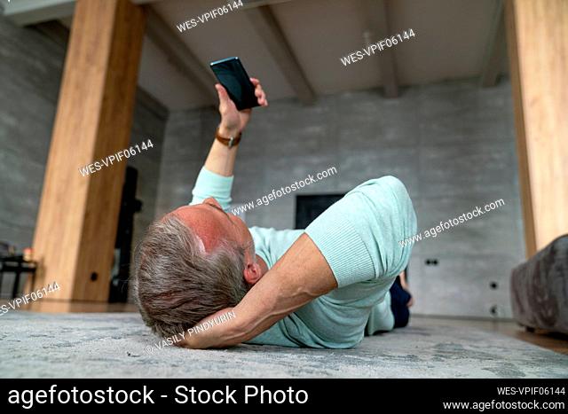 Mature man with hand behind head using smart phone lying on carpet at home