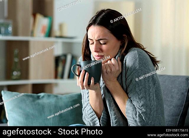 Angry woman getting cold holding coffee mug and grabbing sweater sitting on a couch at home