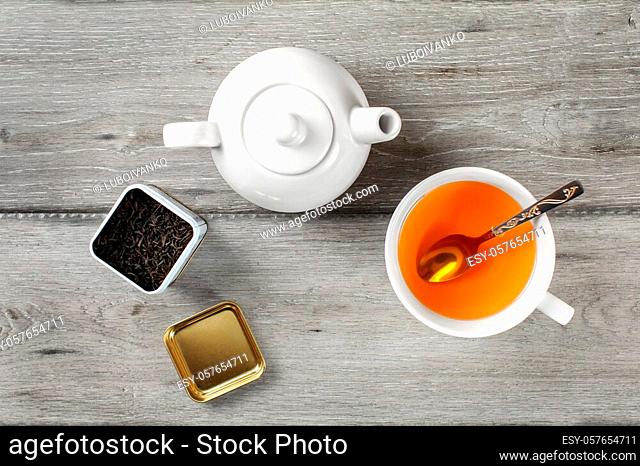 Table top view - brass tea caddy, white porcelain teapot and ceramic cup with silver spoon and hot amber drink, on gray wood desk