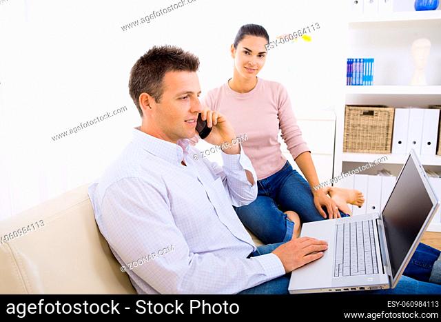 Young couple using laptop computer at home, sitting on couch. Man talkin on mobile phone. Selective focus on man