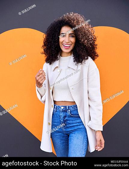Cheerful young woman with curly hair standing against heart shape on wall