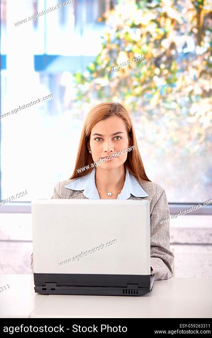 Portrait of confident young businesswoman sitting at desk in office, working on laptop, smiling