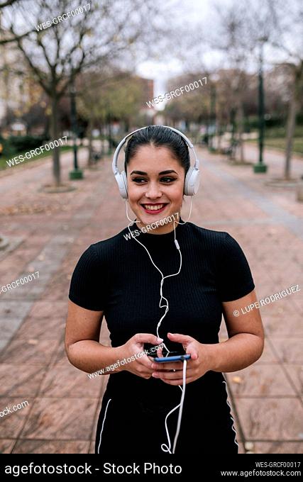 Portrait of happy young woman in black sportswear listening music with headphones and smartphone