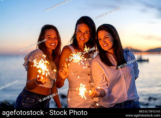 Smiling female friends holding sparklers at beach