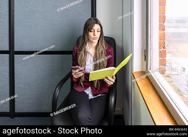 Young blonde business woman sitting in her office using the phone while checking notes