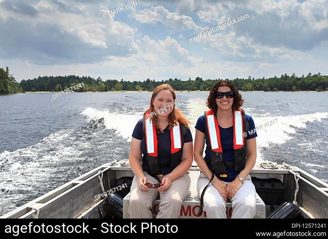Two women in lifejackets sitting in a motorboat looking at the camera as it travels across a lake