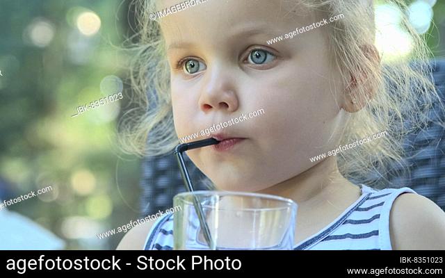 Cute little girl drinks juice through straw. Close-up portrait of blonde girl drinks juice from glass through cocktail straw sitting in street cafe on the park