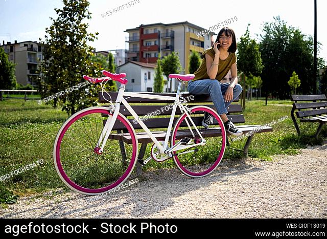 Young woman with with bicycle looking away talking on smart phone while sitting on bench