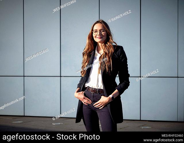 Smiling businesswoman standing with hands in pockets against wall