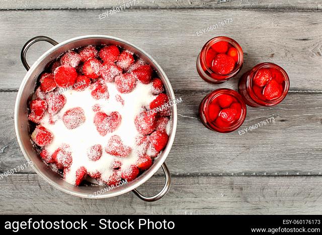 Tabletop view - large steel pot with strawberries covered in crystal sugar, three pickled strawberry glass bottles next to it