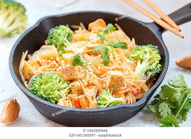 Fried vegan rice noodles with tofu and broccoli in cast iron frying pan, light background. Healthy vegan food concept