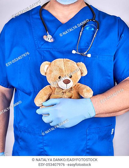 doctor in blue uniform and old latex gloves holding a brown teddy bear, gray background