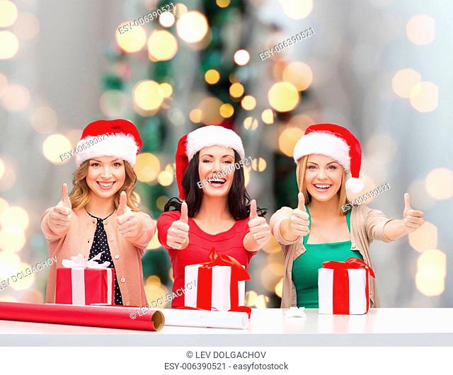 holidays, gesture, decoration and people concept - smiling women in santa helper hats with decorating paper and gift boxes showing thumbs up over christmas tree...