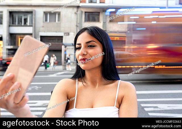 Young woman taking selfie through smart phone on footpath in city