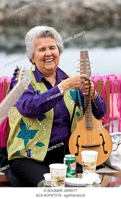 A smiling elder first nations senior adjusts her mandolin during a musical gathering of first nations friends on the deck of a restaurant in Alert Bay