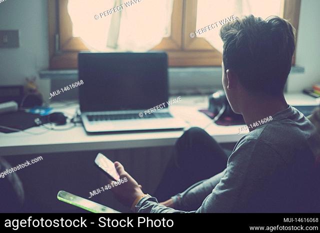 one young man using phone and looking at the laptop or computer at home in his room playing video games. Teenager addicted at social network and social media