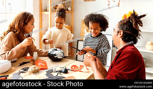 Young joyful african american family mother, father and two kids preparing for Halloween at home, painting pumpkins and making paper cuttings and decorations...