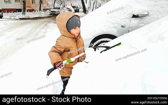 Portrait of charming smiling boy in gloves hat and jacket helping to clean up snow covered parents car after blizzard using big brush