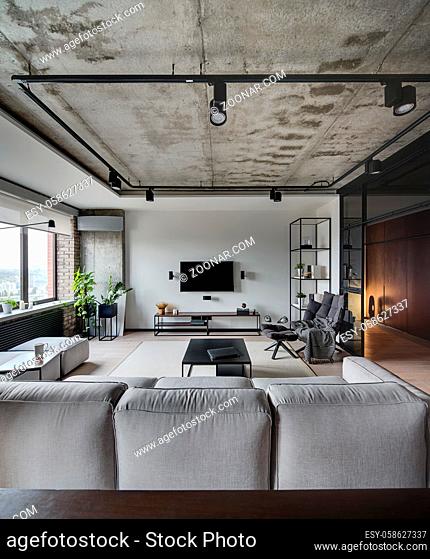 Loft style sitting-room with white, brick and concrete walls. There is sofa, tables, chair, armchair, shelves, speakers, TV and rack under it, lamps
