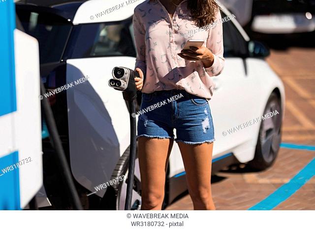 Woman using mobile phone while holding charging plug at charging station
