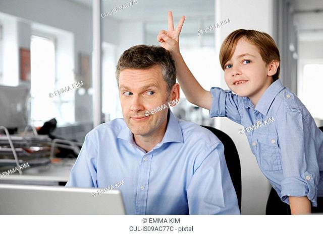 Father using laptop, son making peace sign behind his head