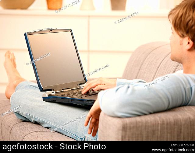 Man using laptop computer sitting on sofa in living room with feet up