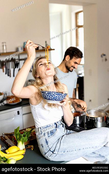 Woman slurping noodles with boyfriend preparing food in kitchen at home